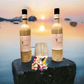 Two bottles of Loudyvie's Kremas, a cream liqueur from Loudyvie's, stand on a stone ledge with a small, empty glass between them. A colorful flower lies in front of the bottles. The background features a serene, blurry sunset over a body of water, adding to the tranquil ambiance of this coconut drink scene.