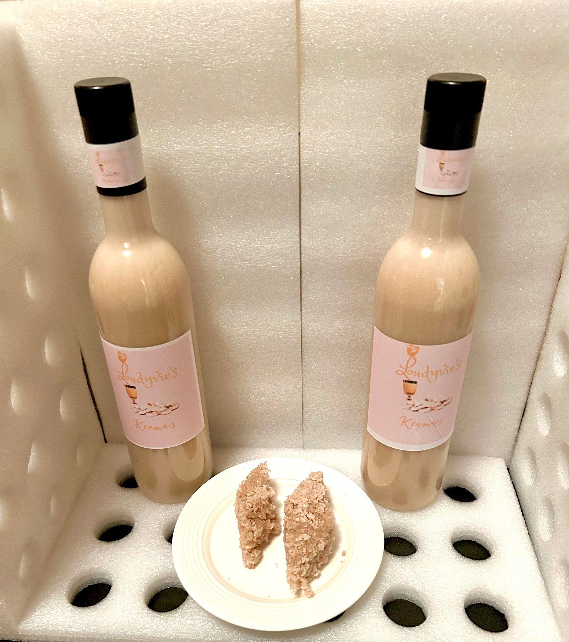 Displayed in a foam-padded box are two bottles of Loudyvie’s Kremas by Loudyvie's. In front of the bottles, a white plate holds two triangular slices of homemade eggnog dessert. The setup highlights the Kremas and the dessert within a secure, cushioned environment.